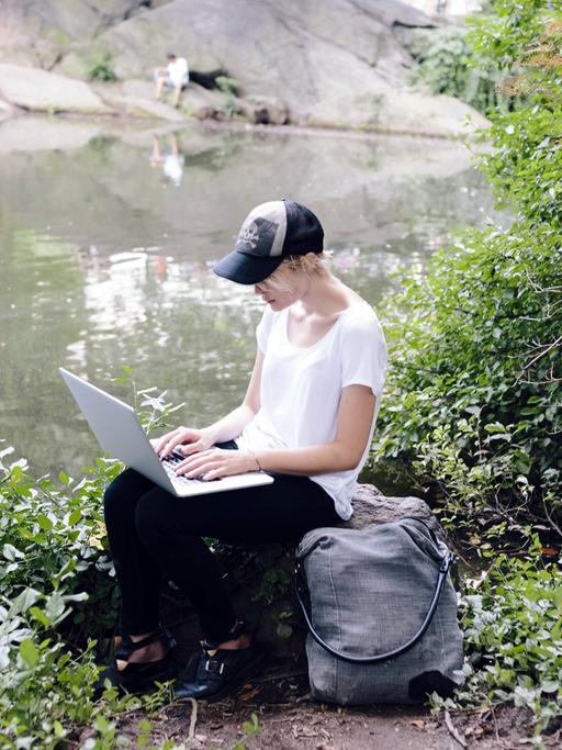 Eine junge Frau sitzt mit ihrem Laptop an einem Seeufer im Grünen