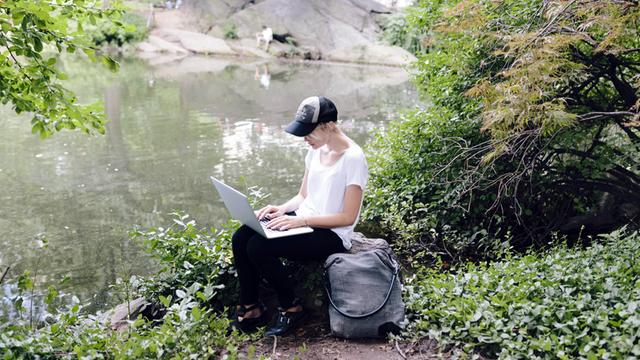 Eine junge Frau sitzt mit ihrem Laptop an einem Seeufer im Grünen