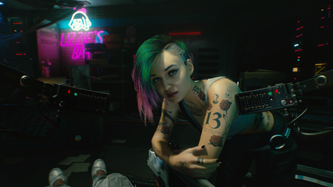 Screenshot aus dem Rollenspiel "Cyberpunk 2077:Eine junge Frau mit vielen Tattoos und bunten, auf einer Seite abrasierten Haaren beugt sich zum Betrachter und blickt ihn ernst an. Hinter ihr befindet sich der Neon-Schriftzug "Lizzie's Bar"