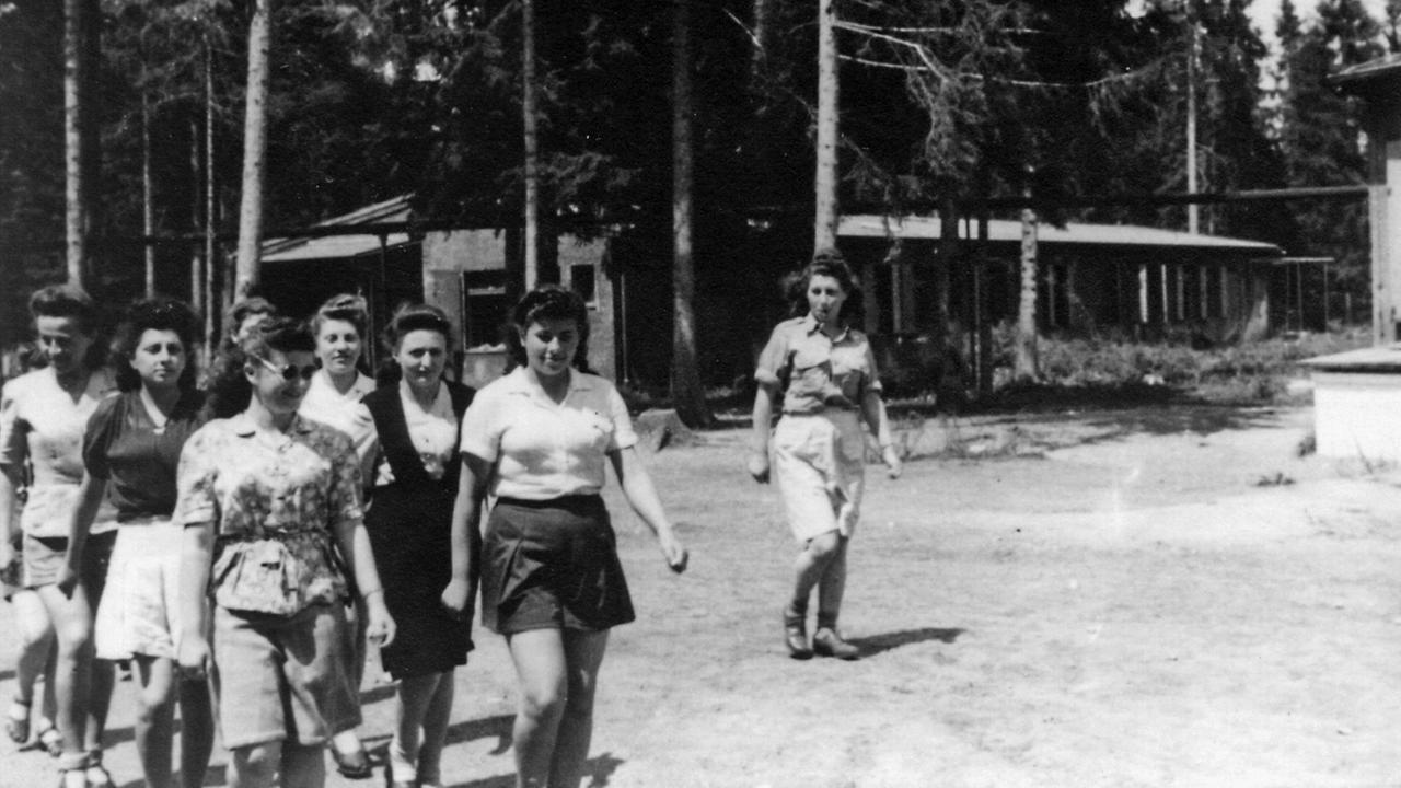Eine Gruppe Frauen in Zivilkleidung läuft, im Hntergrund Baracken zwischen Bäumen