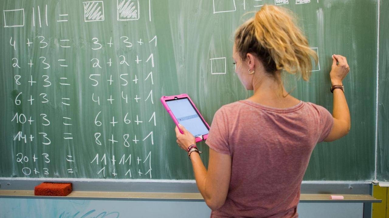 Eine Lehrerin steht mit einem Tablet in der Hand an der Tafel und schreibt Mathe-Aufgaben an Eine Lehrerin steht mit einem Tablet in der Hand an der Tafel und schreibt Mathe-Aufgaben an