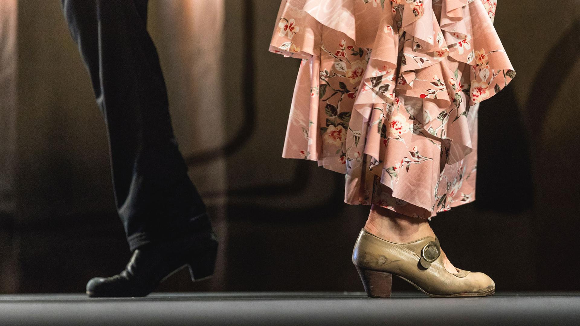  Die Beine von zwei Flamenco-Tänzern (Mann und Frau) sind bis zu den Knien zu sehen. Der Mann trägt eine schwarze Hose, die Frau ein rosafarbenes gestuftes Kleid bis zu den Knöcheln.