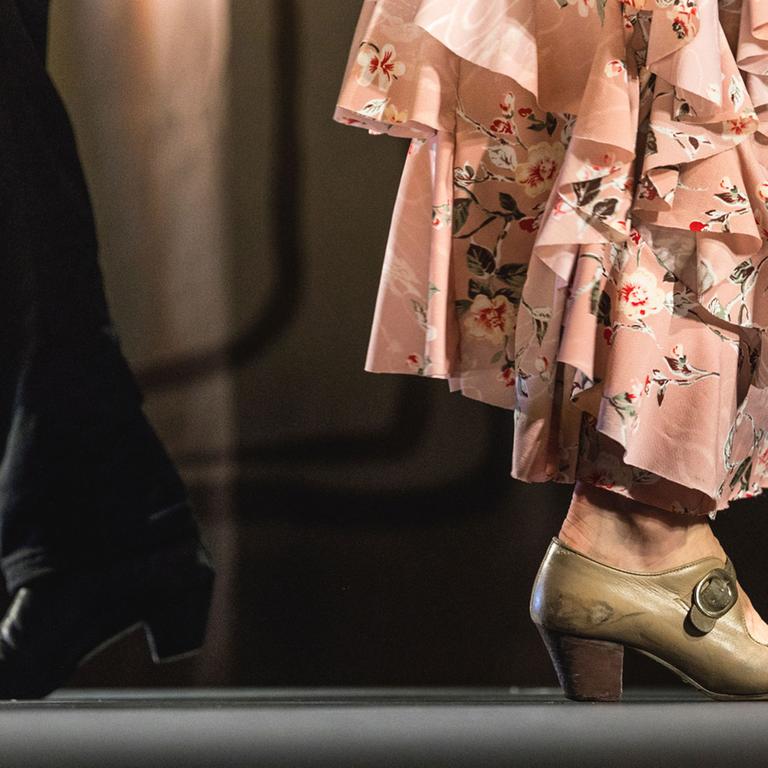 Die Beine von zwei Flamenco-Tänzern (Mann und Frau) sind bis zu den Knien zu sehen. Der Mann trägt eine schwarze Hose, die Frau ein rosa, gestuftes Klein bis zu den Knöcheln. 