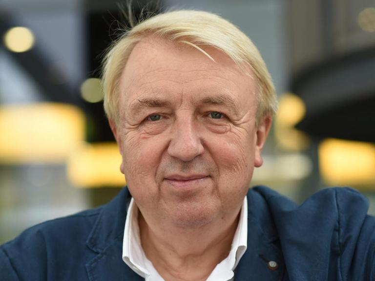 Hanns-Josef Ortheil hat blonde Haar und trägt ein weißes Hemd und dunkle Jacke. Er blickt in Richtung Kamera
