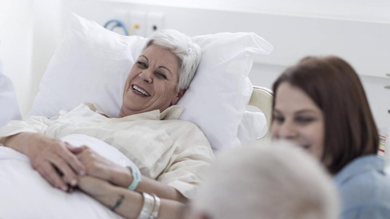 Symbolfoto: Eine weibliche Patientin im Krankenhausbett, rechts Besucher am Bett, links ein Arzt. Die Patientin lacht.