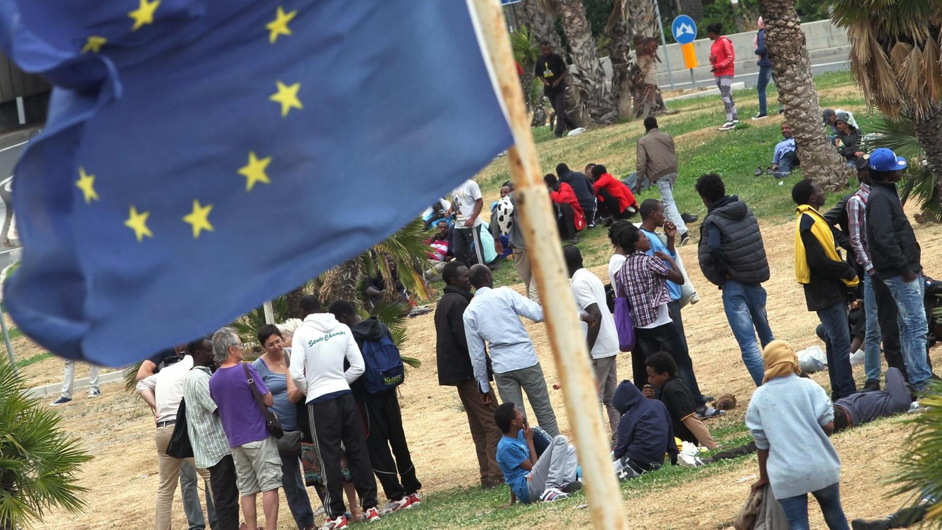 Asylbewerber stehen zusammen, im Vordergrund weht eine EU-Flagge Asylbewerber stehen zusammen, im Vordergrund weht eine EU-Flagge