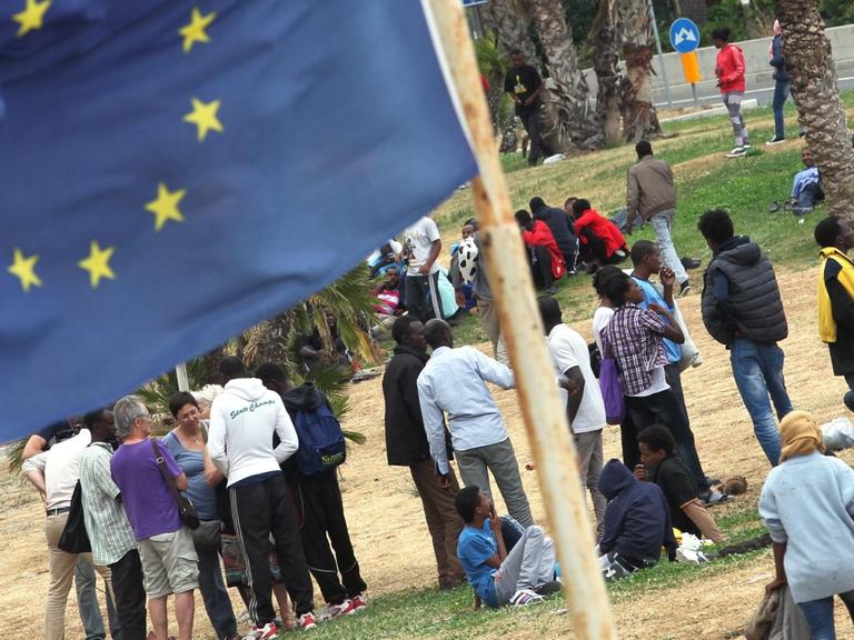 Asylbewerber stehen zusammen, im Vordergrund weht eine EU-Flagge