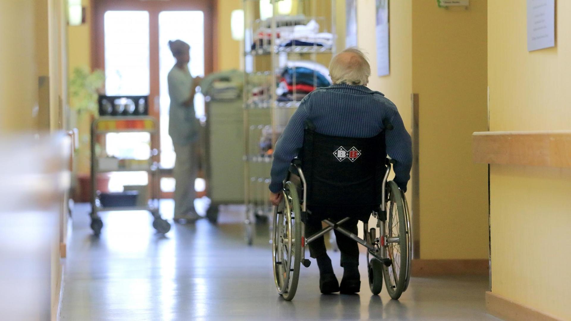 Ein Rentner fährt mit seinem Rollstuhl über einen Flur. Im Hintergrund ist eine Pflegekraft beschäftigt. Ein Rentner fährt mit seinem Rollstuhl über einen Flur. Im Hintergrund ist eine Pflegekraft beschäftigt.