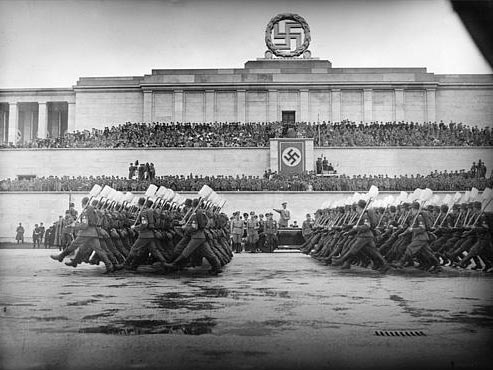 Eine Parade des Reichsarbeitsdienstes: Männer in Uniform und mit Spaten laufen in Reihen vor dem Reichsparteitagsgelände vorbei, auf der Tribüne steht Hitler. 