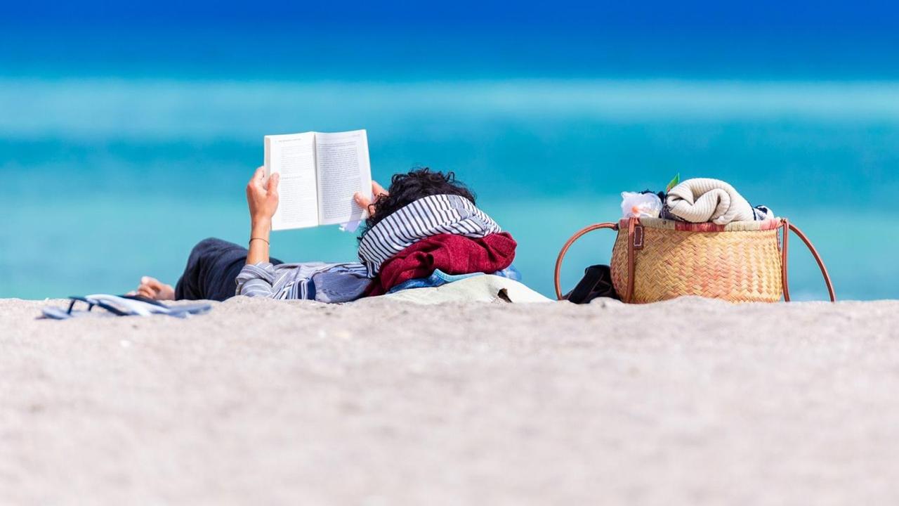 Eine Person liegt vor dem Meer am Strand und liest ein Buch