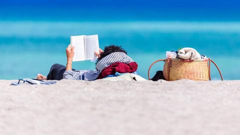 Eine Person liegt vor dem Meer am Strand und liest ein Buch