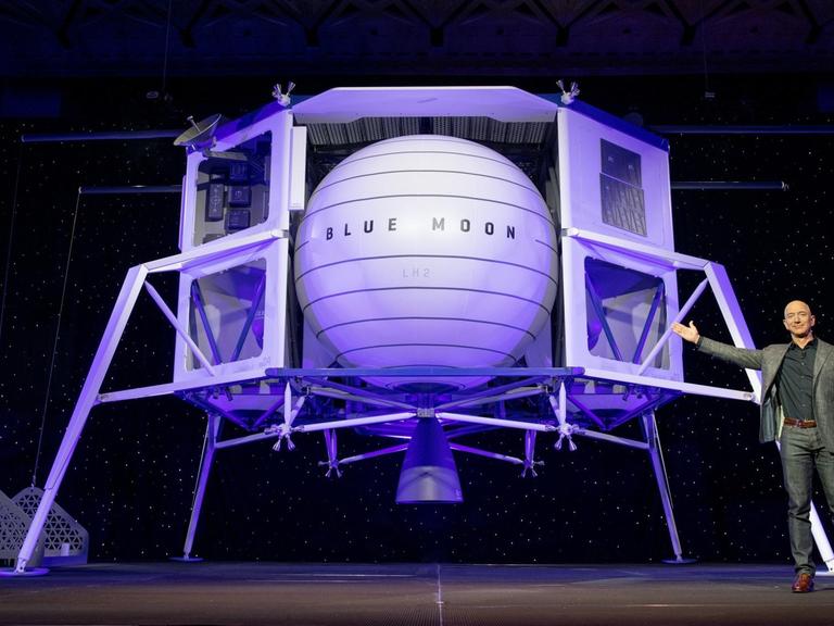 Amazon-Gründer Jeff Bezos steht auf ener Bühne und präsentiert hinter ihm eine in hellem lila angestrahlte Mondlandefähre.