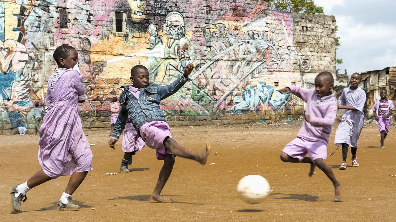 Schulkinder spielen Fußball auf dem sandigen Boden im Mathare Slum. Im Hintergrund ist einer Wand mit Graffiti zu sehen.

