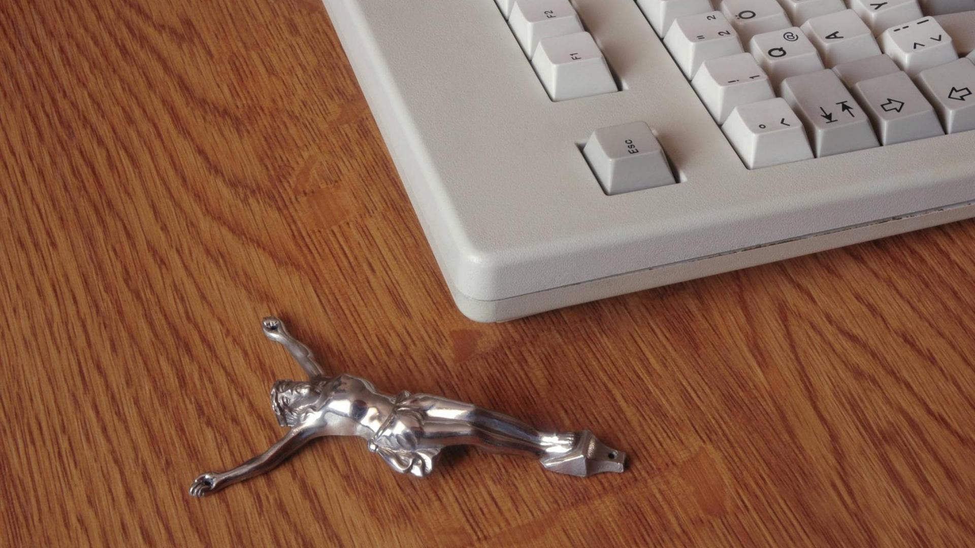 Eine eiserne Jesusfigur liegt auf einem Holztisch neben einer in die Jahre gekommenden Computertastatur. Eine eiserne Jesusfigur liegt auf einem Holztisch neben einer in die Jahre gekommenden Computertastatur.