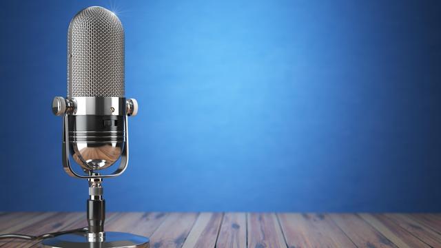 Vor einem blauen Hintergrund steht auf einem Holzboden ein Retro-Mikrofon aus silber-glänzendem Material.