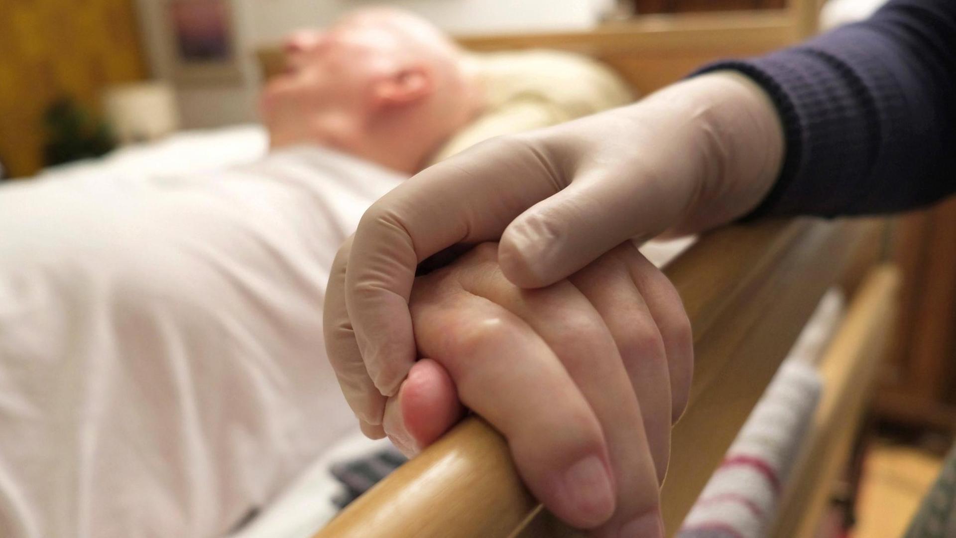 Die Hand einer Altenpflegerin in Schutzhandschuh hält die Hand eines alten Menschen in einem Seniorenheim. Die Hand einer Altenpflegerin in Schutzhandschuh hält die Hand eines alten Menschen in einem Seniorenheim.