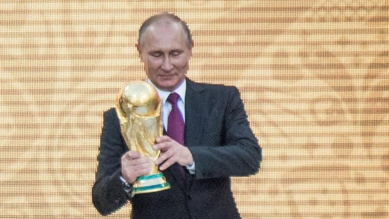 Putin (re.) hält eine goldene Trophäe, Infantino kommt von links auf ihn zu. Vor der Bühne stehen Zuschauer. Putin (re.) hält eine goldene Trophäe, Infantino kommt von links auf ihn zu. Vor der Bühne stehen Zuschauer.
