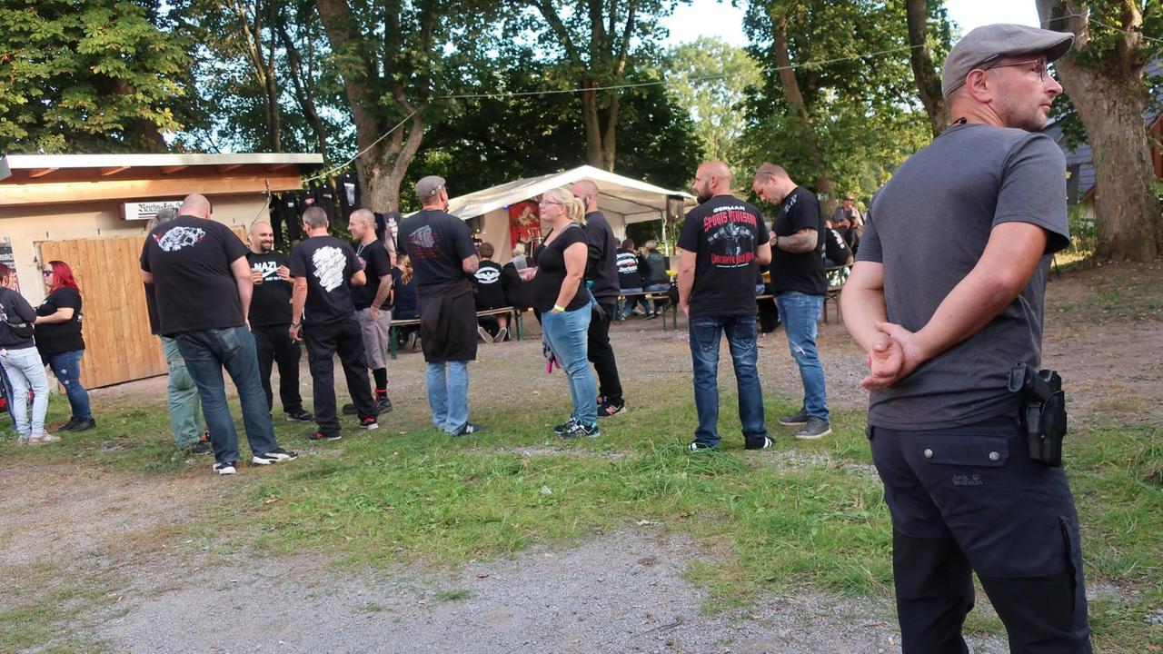 Im Vordergrund steht ein bewaffneter Polizist in Zivil, im Hintergrund stehen weibliche und männliche Neonazis in einem Garten Im Vordergrund steht ein bewaffneter Polizist in Zivil, im Hintergrund stehen weibliche und männliche Neonazis in einem Garten