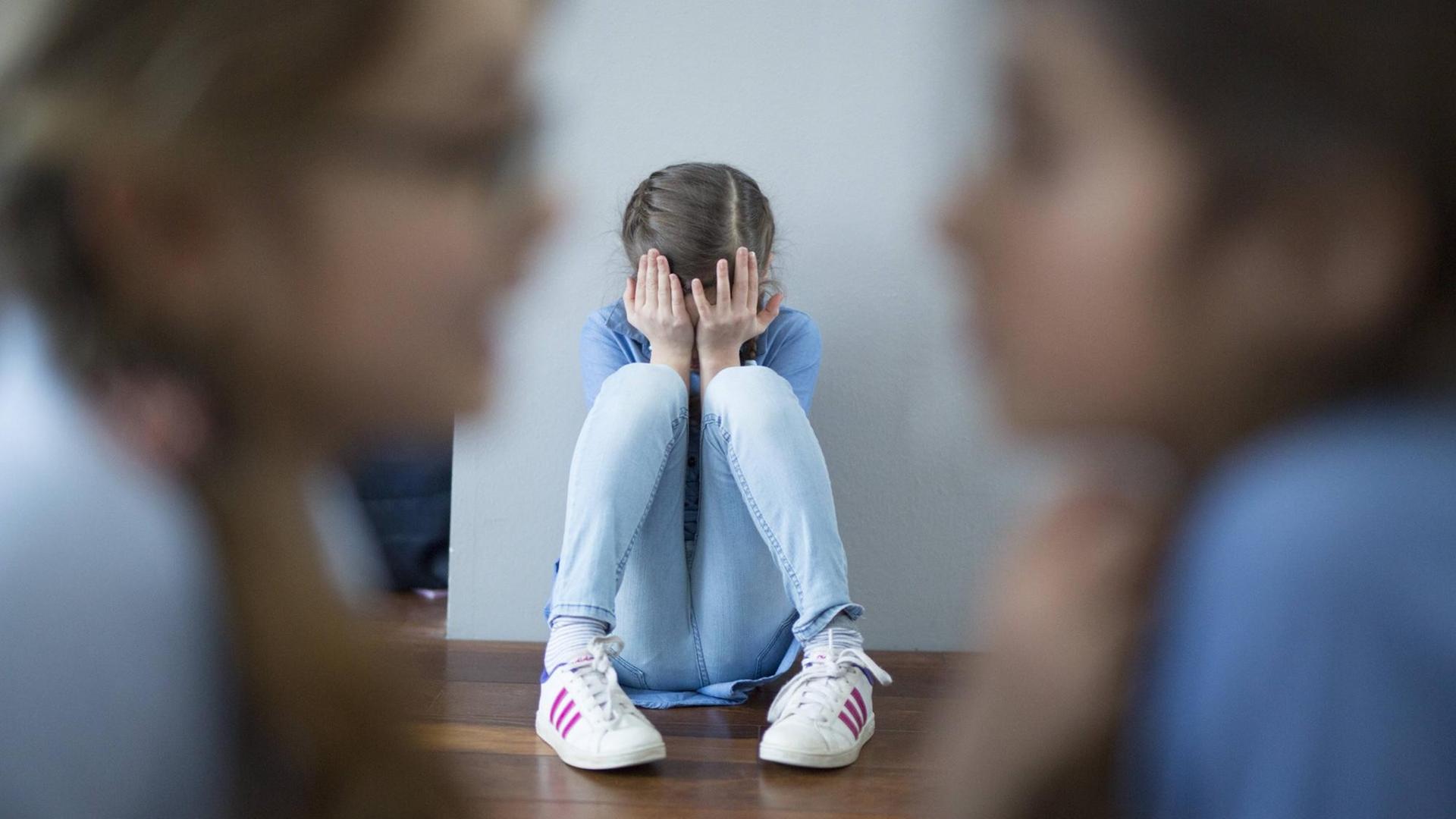 Zwei Mädchen lästern über ein drittes, das an der Wand kauert und die Hände vors Gesicht geschlagen hat. Zwei Mädchen lästern über ein drittes, das an der Wand kauert und die Hände vors Gesicht geschlagen hat.
