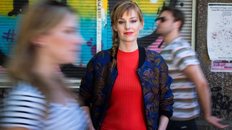 Eva Schulz vor buntem Hintergrund. Vor und hinter ihr gehen Menchen vorbei. Eva Schulz vor buntem Hintergrund. Vor und hinter ihr gehen Menchen vorbei.