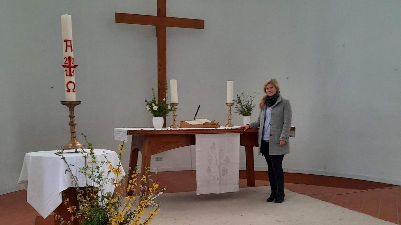 Ein blonde Frau in grauer Jacke und dunklen Hosen steht in einem Kirchenschiff. An der Wand hängt ein großes schlichtes Holzkreuz.