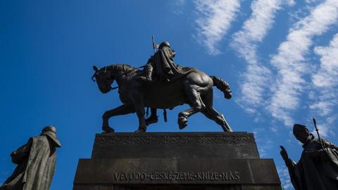 Reiterdenkmal des Hl. Wenzel: die Wenzelsstatue vor dem Nationalmuseum in Prag, der seine Lanze in den blauen Himmel reckt und dem eine geistliche Bronze-Person folgt.