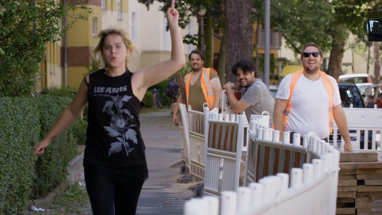 Eine Frau geht wild gestikulierend über eine Straße, während Bauarbeiter an einer Baustelle lachen. Eine Frau geht wild gestikulierend über eine Straße, während Bauarbeiter an einer Baustelle lachen.
