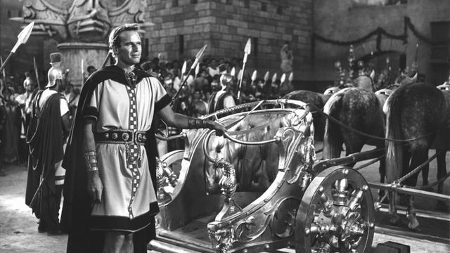 Ein Mann in römischer Uniform steht neben einem Pferderennwagen und hält die Zügel in die Hand.