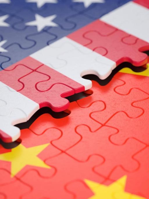 Symbolbild: Ein unfertiges Puzzle, das einen Teil der Flagge der USA zeigt, liegt über einem anderen Puzzle, das einen Teil der Flagge Chinas zeigt.