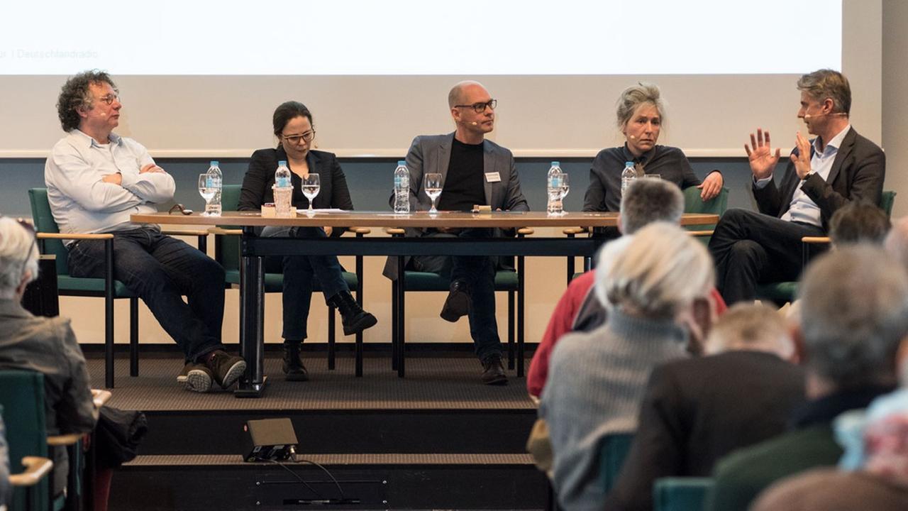 Auf einer Bühne sitzen fünf Gesprächsteilnehmerinnen. Im Vordergrund ist Publikum zu sehen. Auf einer Bühne sitzen fünf Gesprächsteilnehmerinnen. Im Vordergrund ist Publikum zu sehen.