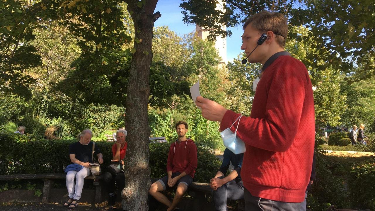 Theaterprojekt "Kein Schlussstrich!" - Stadtrundgang, Menschen sitzen auf zwei Parkbänken. Im Fordergrund ein junger Mann mit Kopghörer und Mikrophon, einen Zettel in der Hand haltend. Theaterprojekt "Kein Schlussstrich!" - Stadtrundgang, Menschen sitzen auf zwei Parkbänken. Im Fordergrund ein junger Mann mit Kopghörer und Mikrophon, einen Zettel in der Hand haltend.