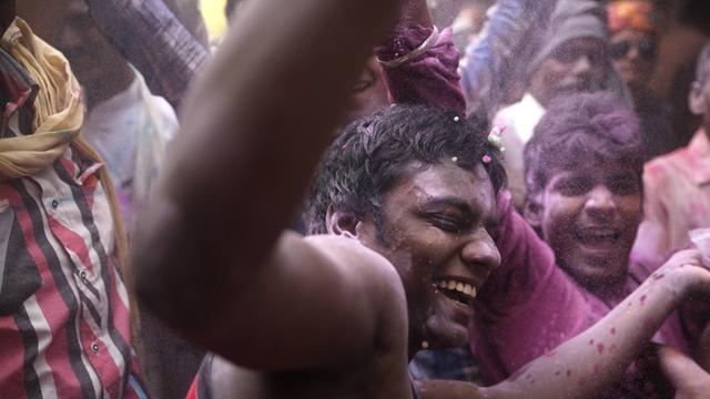 Ein Teenager tanzt und lacht auf dem Holi Spring Festival of Colours in Uttar Pradesh, Mathura in Indien. Im Hintergrund sind viele andere Tänzer, die alle mit pinken Farbpulver bedeckt sind.