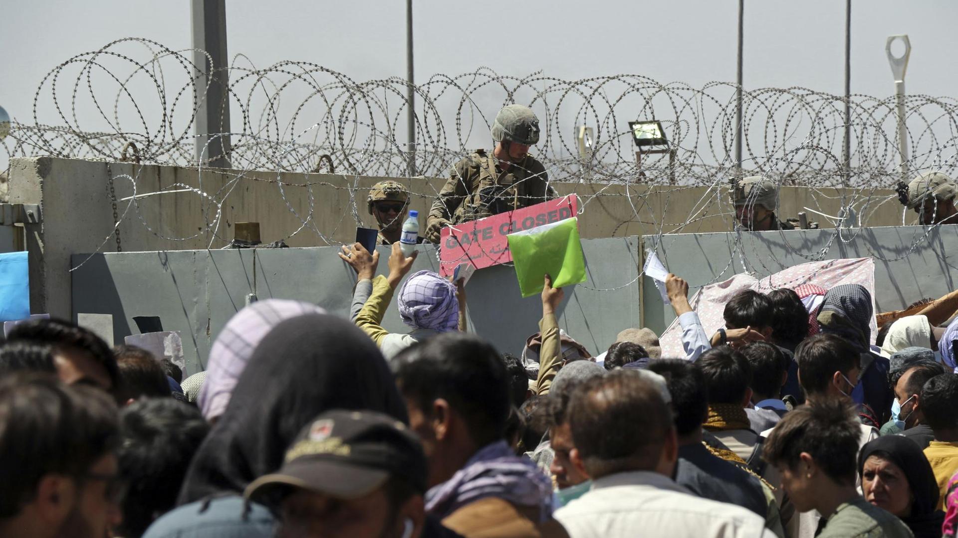 Ein US-Soldat steht hinter einem Stacheldrahtzaun, davor stehen viele Menschen und halten ihre Papiere in die Höhe.