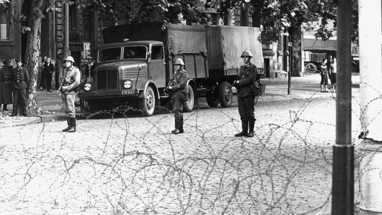 Hinter dem Stacheldrahtverhau in der Bernauer Straße stehen Volkspolizisten mit dem Gewehr im Anschlag. Am frühen Sonntagmorgen des 13. August 1961 wurde unter der Aufsicht von bewaffneten Streitkräften der DDR mit der Errichtung von Straßensperren aus Stacheldraht und dem Bau einer Mauer begonnen, um den Ostteil Berlins vom Westteil abzusperren. Die Mauer soll den ständig steigenden Flüchtlingsstrom von Ost- nach West-Berlin stoppen. Hinter dem Stacheldrahtverhau in der Bernauer Straße stehen Volkspolizisten mit dem Gewehr im Anschlag. Am frühen Sonntagmorgen des 13. August 1961 wurde unter der Aufsicht von bewaffneten Streitkräften der DDR mit der Errichtung von Straßensperren aus Stacheldraht und dem Bau einer Mauer begonnen, um den Ostteil Berlins vom Westteil abzusperren. Die Mauer soll den ständig steigenden Flüchtlingsstrom von Ost- nach West-Berlin stoppen.
