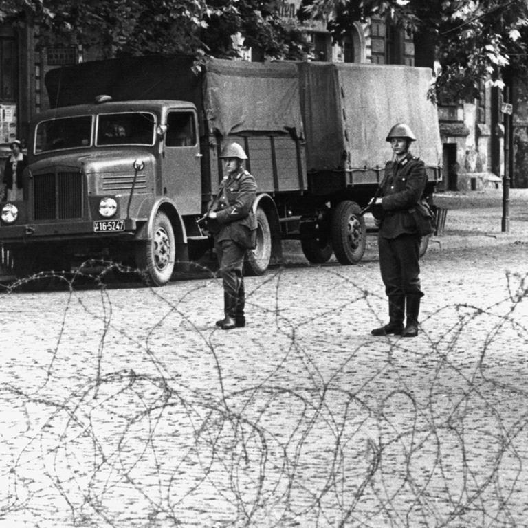 Hinter dem Stacheldrahtverhau in der Bernauer Straße stehen Volkspolizisten mit dem Gewehr im Anschlag. Am frühen Sonntagmorgen des 13. August 1961 wurde unter der Aufsicht von bewaffneten Streitkräften der DDR mit der Errichtung von Straßensperren aus Stacheldraht und dem Bau einer Mauer begonnen, um den Ostteil Berlins vom Westteil abzusperren. Die Mauer soll den ständig steigenden Flüchtlingsstrom von Ost- nach West-Berlin stoppen.