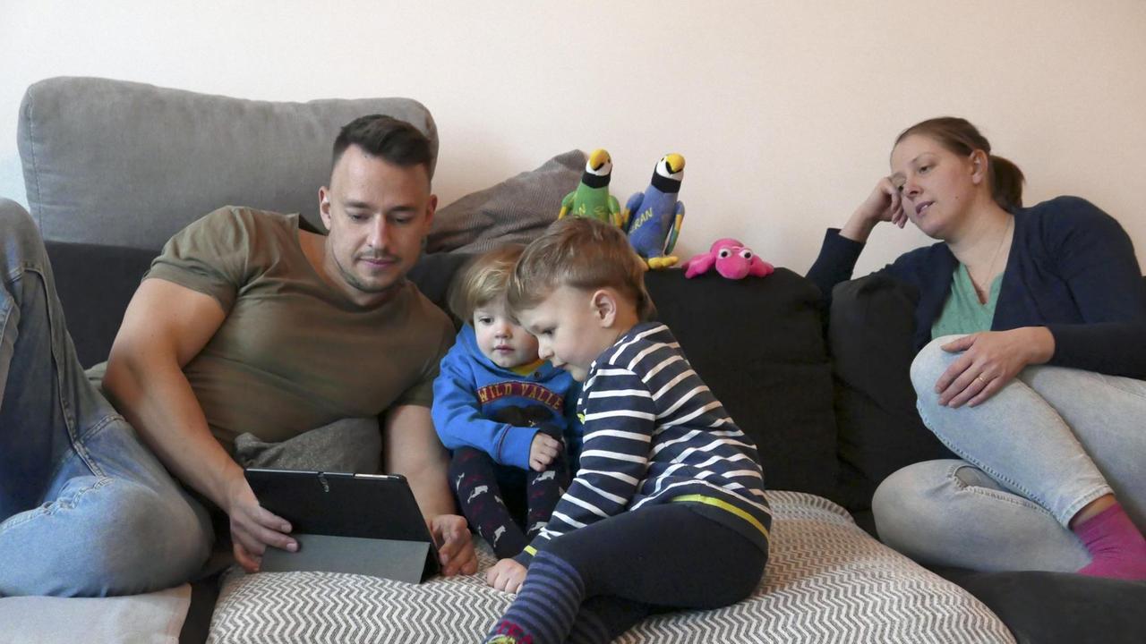 Ein junger Mann und seine zwei Kinder sitzen auf der auf der Couch und schauen auf ein Tablet, eine junge Frau sitzt rechts neben ihnen. Ein junger Mann und seine zwei Kinder sitzen auf der auf der Couch und schauen auf ein Tablet, eine junge Frau sitzt rechts neben ihnen.