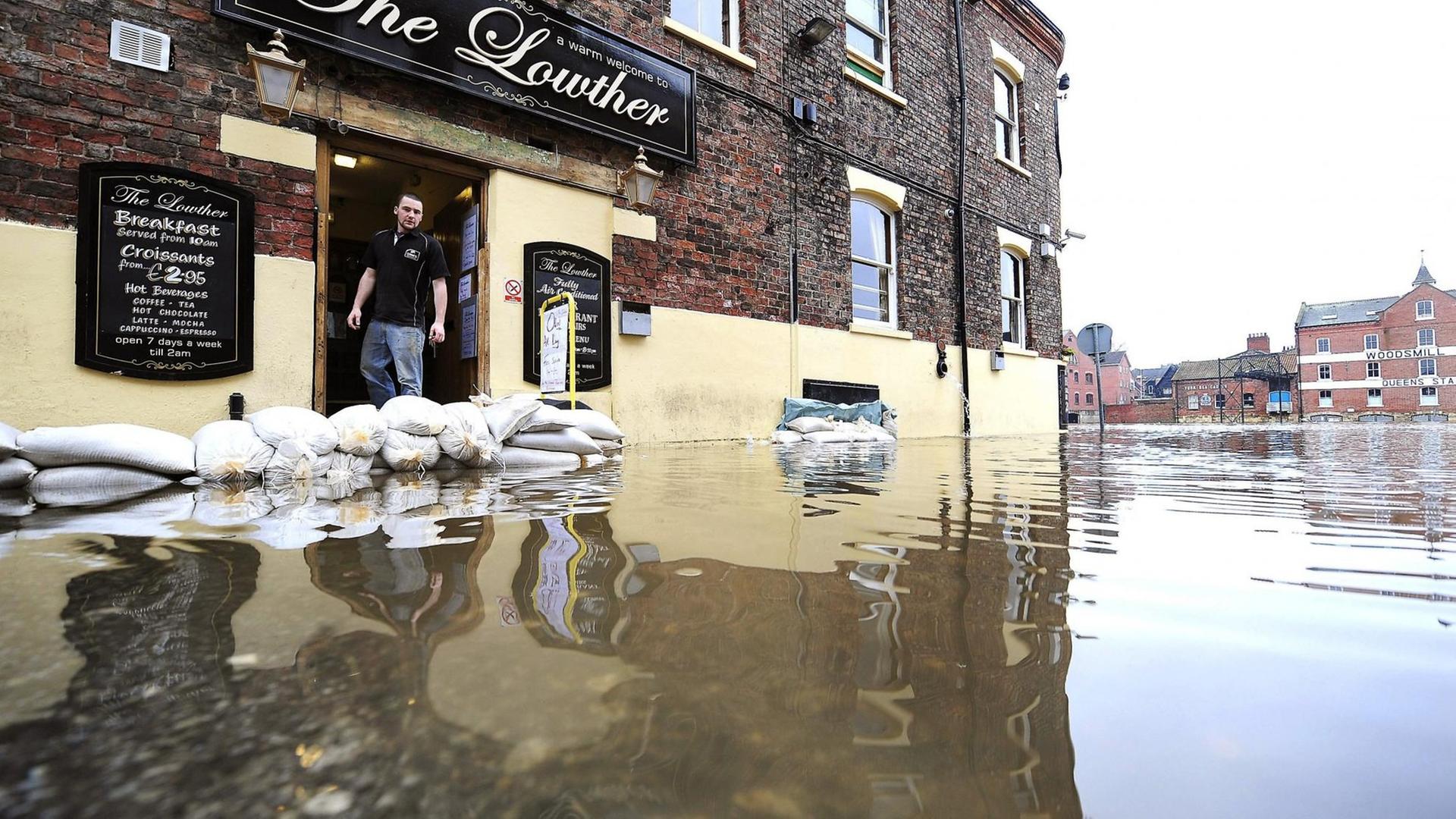 Eine Straße, steht durch Überschwemmungen völlig unter Wasser. Auf der linken Seite  steht ein Mann in der Tür seines Pubs.