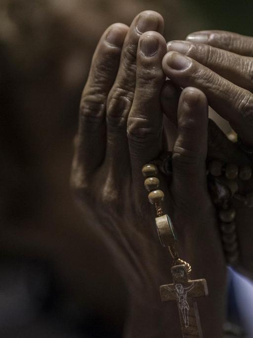 Dunkelhäufige Hände halten beim Gebet eine Kette mit einem Kreuz.