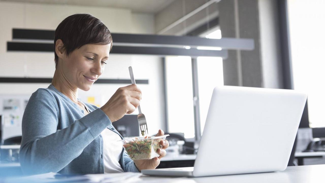 Eine junge Frau isst einen Salat vor ihrem aufgeklappten Laptop.