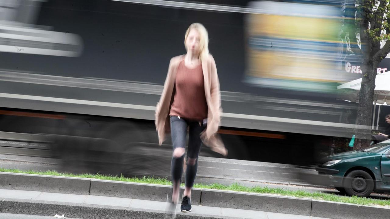 Junge Frau mit langen blonden Haaren steht am Straßenrand mit schnell an ihr vorbeifahrenden Autos. 