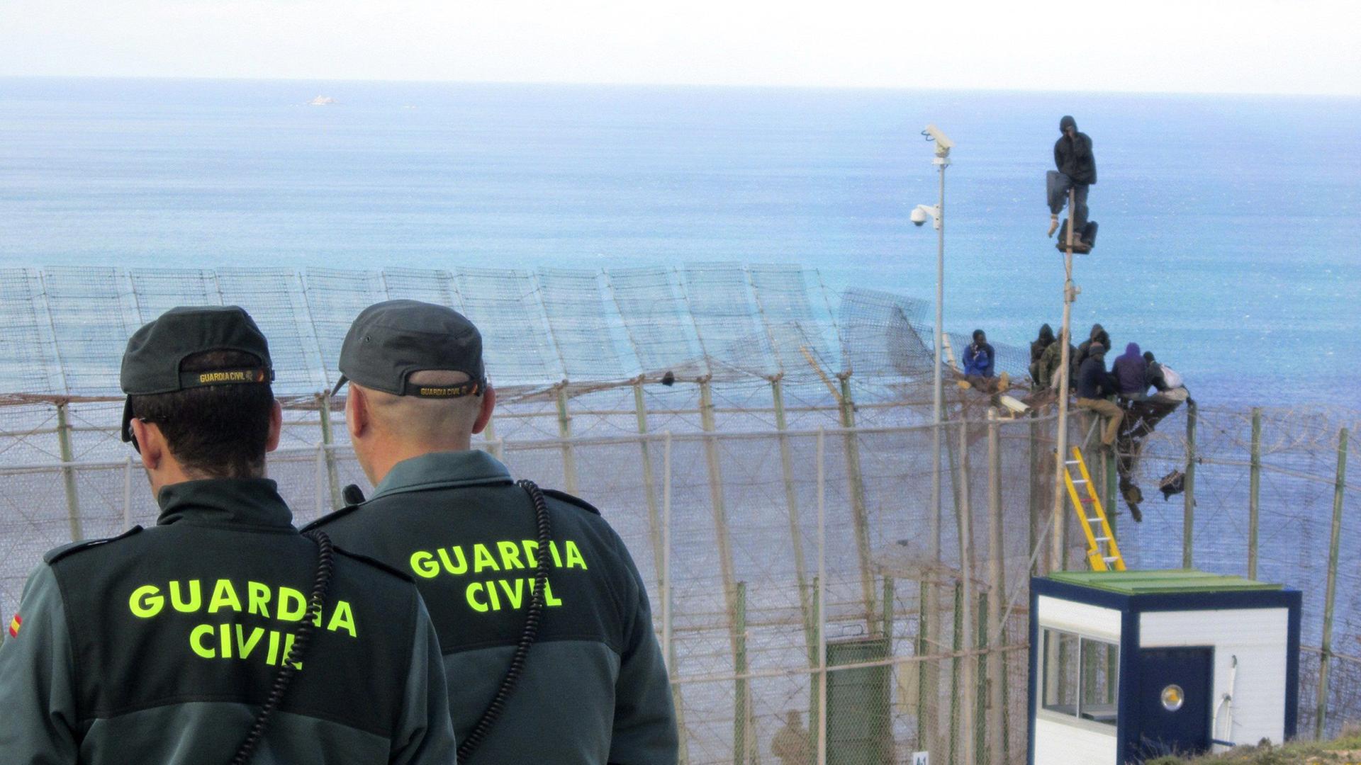 Polizisten der spanischen Guardia Civil beobachten, wie afrikanische Flüchtlinge den Grenzzaun zur spanischen Exklave Melilla überwinden Polizisten der spanischen Guardia Civil beobachten, wie afrikanische Flüchtlinge den Grenzzaun zur spanischen Exklave Melilla überwinden
