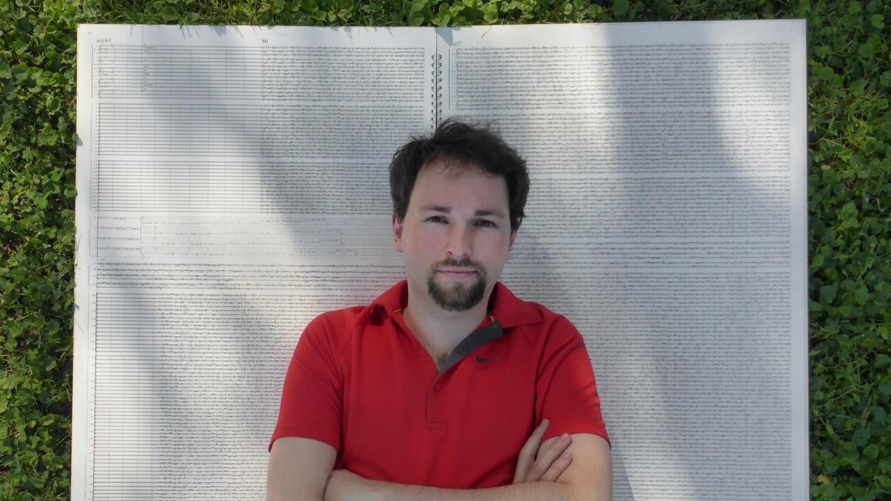 Ein junger Mann, von oben fotografiert, liegt auf einer Kopfkissen-großen Partitur, die auf einem Rasenstück aufgeschlagen ist. Ein junger Mann, von oben fotografiert, liegt auf einer Kopfkissen-großen Partitur, die auf einem Rasenstück aufgeschlagen ist.