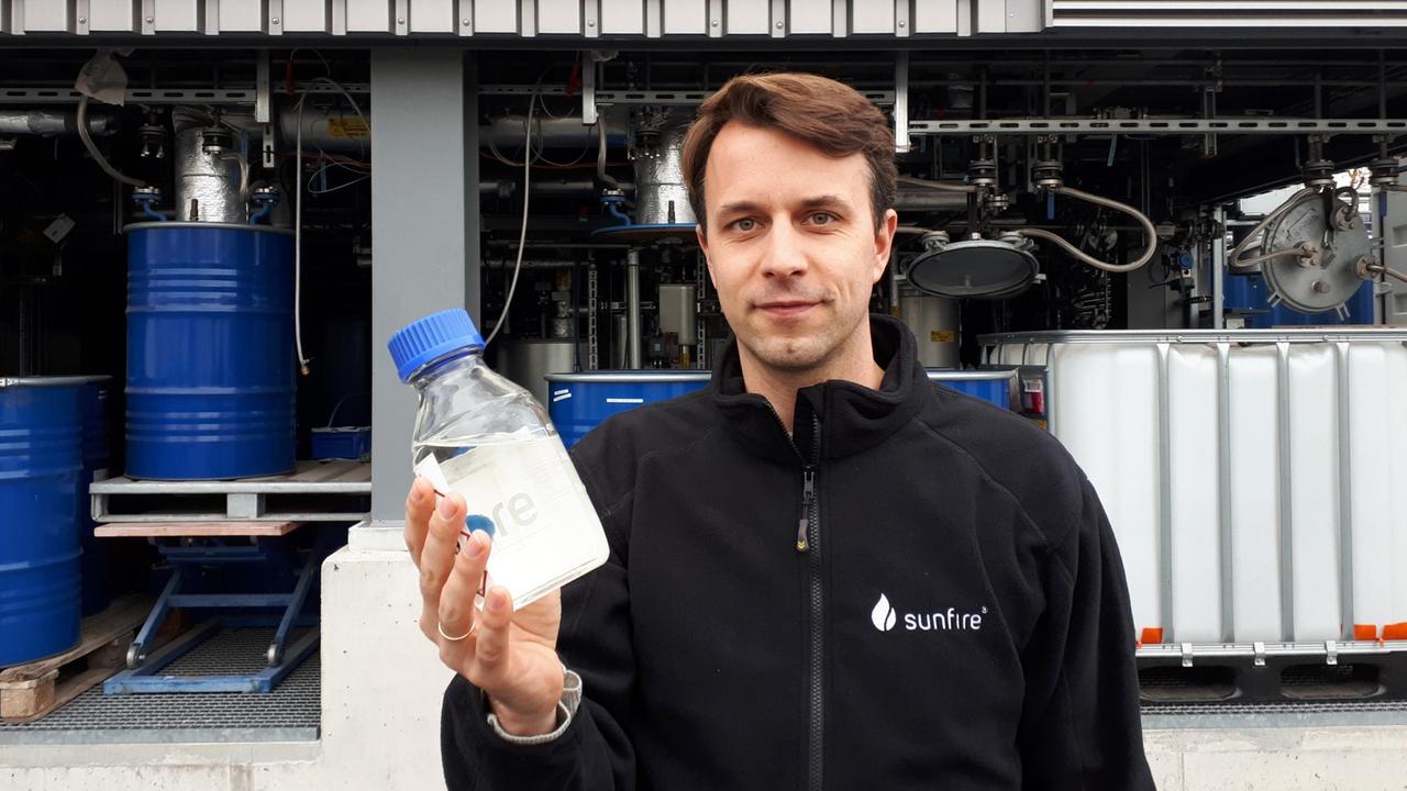 Nils Aldag, Geschäftsführer und Miteigentümer des Dresdener Unternehmens Sunfire. Ein Mann steht auf einem Betriebshof und hält eine Flasche mit einer durchsichtigen Flüssigkeit in der Hand.