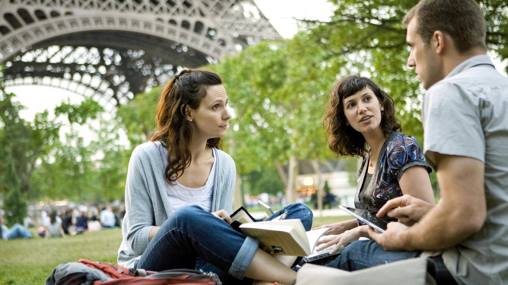 Zwei Frauen und ein Mann mit Bücher und Tablet in der Hand im Gespräch auf einer Wiese vor dem Eiffelturm in Paris Zwei Frauen und ein Mann mit Bücher und Tablet in der Hand im Gespräch auf einer Wiese vor dem Eiffelturm in Paris