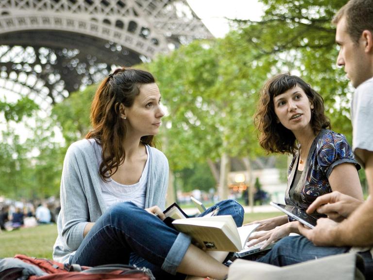 Zwei Frauen und ein Mann mit Bücher und Tablet in der Hand im Gespräch auf einer Wiese vor dem Eiffelturm in Paris
