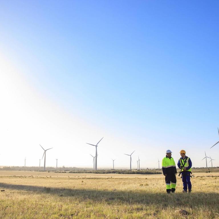 Zwei Männer in Arbeitskleidung stehen auf einer Wiese vor einer Windkraftanlage. Symbolfoto.