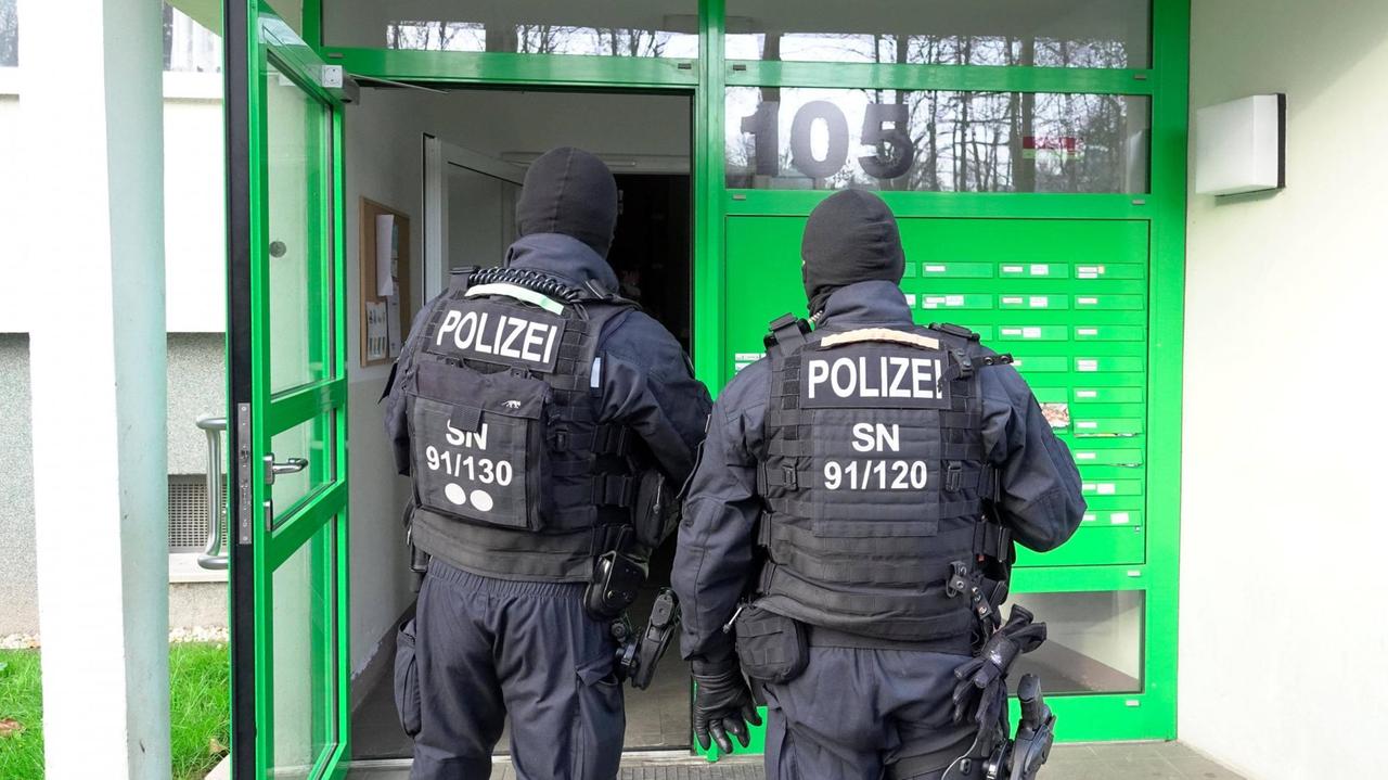 Zwei Polizisten in Kampfmontur stehen vor der grüngestrichenen Eingangstür eines Wohnblocks.
