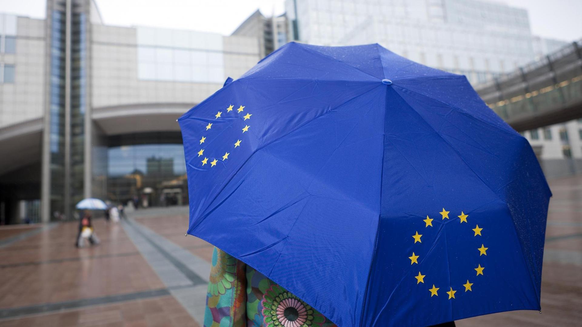 Ein Frau steht im Regen mit einem blauen EU-Schirm vor dem Gebäudekomplex des EU-Parlaments in Brüssel Ein Frau steht im Regen mit einem blauen EU-Schirm vor dem Gebäudekomplex des EU-Parlaments in Brüssel