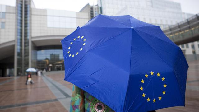 Ein Frau steht im Regen mit einem blauen EU-Schirm vor dem Gebäudekomplex des EU-Parlaments in Brüssel