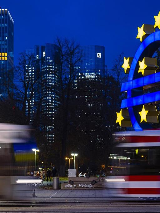 Zwei Straßenbahnen passieren den Theaterplatz in Frankfurt am Main vor dem Euro-Zeichen der Europäischen Zentralbank zur blauen Stunde.